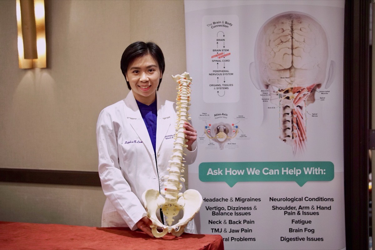 Dr. Sophia B. Lam, Doctor of Chiropractic, in her University of Bridgeport white coat holding a full spine and pelvis model